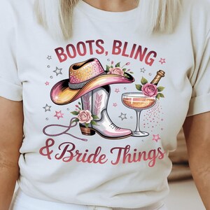 May include: Cream-colored t-shirt with a colorful graphic design featuring a cowboy hat, cowboy boots, a champagne glass, and the text "Boots, Bling & Bride Things". The shirt is paired with white pants.