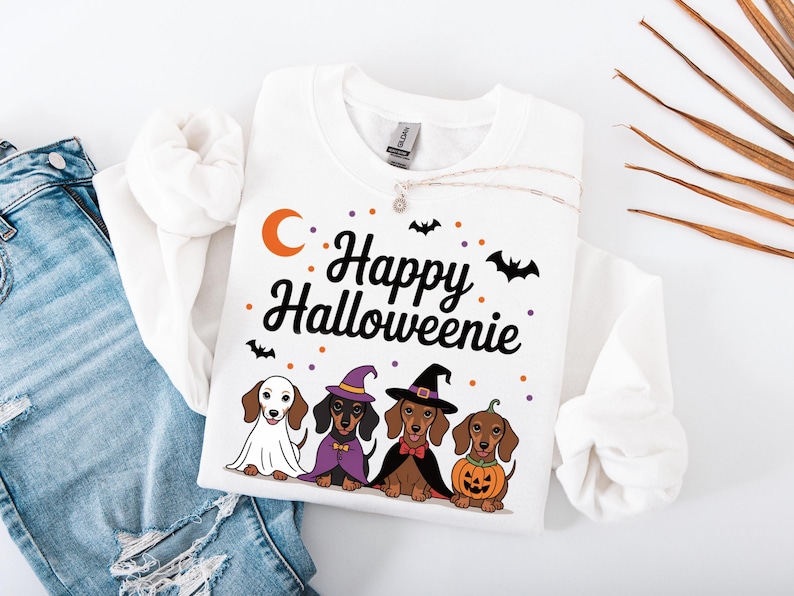 May include: White sweatshirt with the text "Happy Halloweenie" and illustrations of dachshunds in Halloween costumes. The dogs are dressed as a ghost, witch, vampire, and pumpkin. Orange moon and bat graphics.