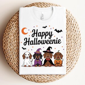 May include: White t-shirt featuring the text "Happy Halloweenie" with a crescent moon and bats. Below the text are four dachshunds dressed in Halloween costumes, including a ghost, witch, and pumpkin.