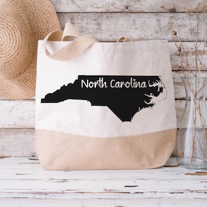 May include: A white canvas tote bag with a black outline of the state of North Carolina and the text "North Carolina" printed on the bag.