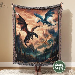 May include: A tapestry throw blanket featuring a fantasy scene with three dragons soaring over a mountainous landscape. The dragons are in shades of black, blue, and gold, set against a backdrop of a sunset sky and forest.