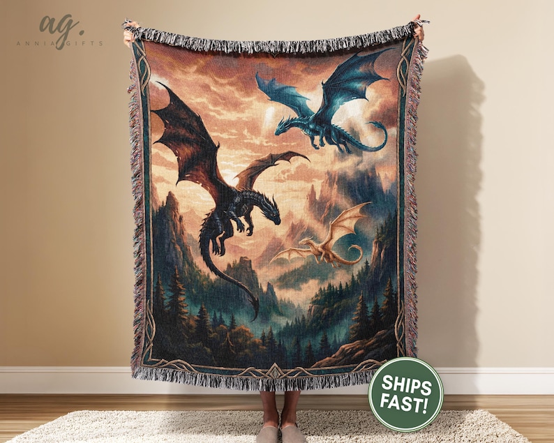 May include: A tapestry throw blanket featuring a fantasy scene with three dragons soaring over a mountainous landscape. The dragons are in shades of black, blue, and gold, set against a backdrop of a sunset sky and forest.