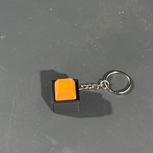 May include: A keychain featuring a square, orange button set in a black frame. The button is attached to a silver chain and a keyring. The background is a solid, gray surface.