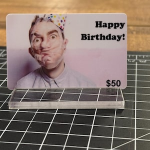Puede incluir: Una tarjeta de regalo blanca con una foto de una persona que lleva un gorro de fiesta sobre un fondo rosa. El texto "Happy Birthday!" est&aacute; impreso en negro en la tarjeta. La tarjeta tambi&eacute;n tiene el texto "$50" impreso en negro.