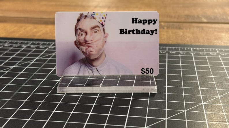 Puede incluir: Una tarjeta de regalo blanca con una foto de una persona que lleva un gorro de fiesta sobre un fondo rosa. El texto "Happy Birthday!" est&aacute; impreso en negro en la tarjeta. La tarjeta tambi&eacute;n tiene el texto "$50" impreso en negro.