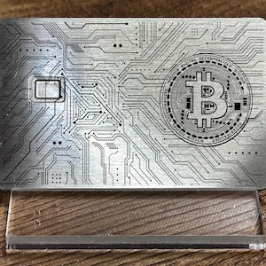 May include: A silver metal card with a circuit board design and a Bitcoin logo in the center. The card is on a clear acrylic stand.