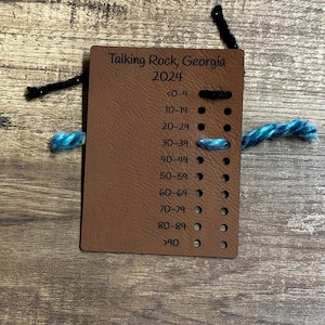 May include: A brown leather card with the text "Talking Rock, Georgia 2024" and a numbered list of age ranges from 0-9 to over 90. There are small black circles next to each age range. A black string and a blue and green string are placed next to the card.