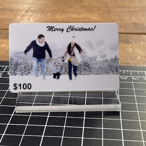 Puede incluir: Una tarjeta de regalo blanca con una foto de una familia en un paisaje invernal nevado. El texto "&iexcl;Feliz Navidad!" est&aacute; impreso en la parte superior de la tarjeta. La tarjeta tiene un valor de 100 &euro;.