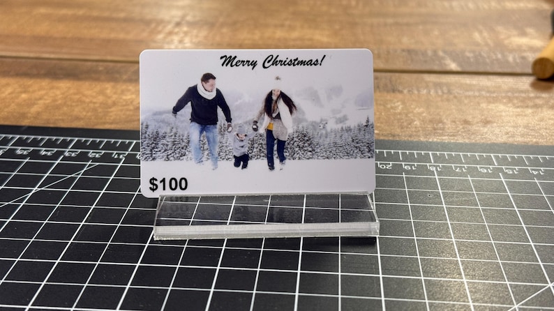 Puede incluir: Una tarjeta de regalo blanca con una foto de una familia en un paisaje invernal nevado. El texto "&iexcl;Feliz Navidad!" est&aacute; impreso en la parte superior de la tarjeta. La tarjeta tiene un valor de 100 &euro;.