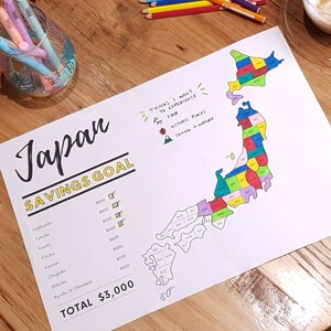 May include: A white sheet of paper with a map of Japan divided into regions. Each region is labelled with a dollar amount and a tick. The text "Japan Savings Goal" is written at the top of the page. The text "Things I Want to Experience" is written at the top right corner of the page, with a list of things to experience in Japan, including food, historic places, and skiing and nature.