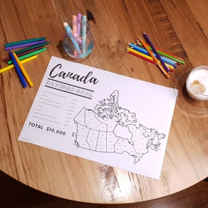 May include: A black and white outline map of Canada with each province and territory labelled with a dollar amount. The map is on a wooden table with coloured pencils and a glass of water. The text "Canada Savings Goal" and "Total $10,000" are at the top of the map.