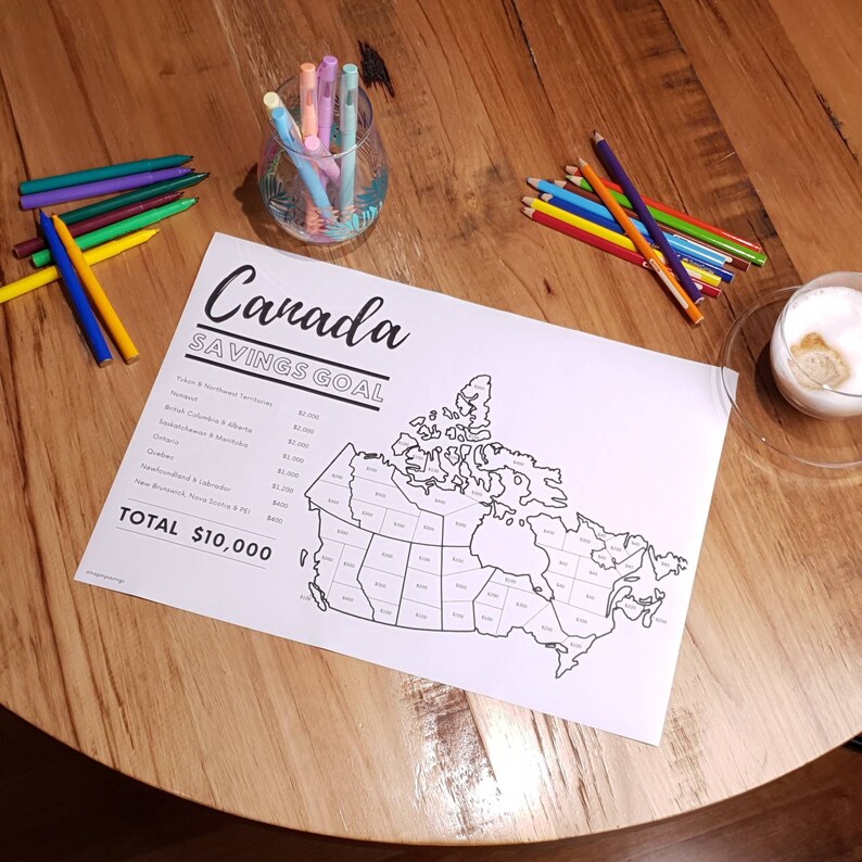 May include: A black and white outline map of Canada with each province and territory labelled with a dollar amount. The map is on a wooden table with coloured pencils and a glass of water. The text "Canada Savings Goal" and "Total $10,000" are at the top of the map.