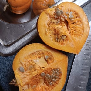 May include: Two halves of a small orange pumpkin with seeds visible inside. The pumpkin is sitting on a stainless steel surface with a white handled knife.