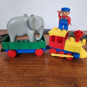 Op de afbeelding: Een kleurrijke plastic speelgoedtreinset met een rode, gele en blauwe locomotief met een klein figuurtje in een blauwe pet en rood shirt bovenop. Er is een groene goederenwagon aangekoppeld met een grote grijze plastic olifant.