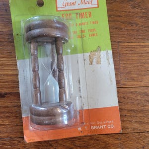 May include: A Grant Maid egg timer in original packaging. The hourglass features a brown wooden frame with white sand inside. The cardboard backing is green and orange with text including Grant Maid Egg Timer and W. T. Grant Co. printed on it.