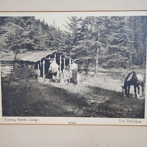 Op de afbeelding: Een zwart-witfoto met het opschrift Jimmy Brook Camp 1942 The Tobique, waarop een rustieke blokhut in een bosrijke omgeving te zien is, met twee personen bij de ingang en een paard dat op de voorgrond bij een beekje graast.