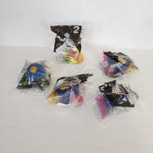 May include: Five sealed clear plastic bags containing small, colorful plastic toys from a McDonald's Happy Meal promotion. Each bag features black printed text and graphics, including the number 2 on one bag, and various toy shapes inside.