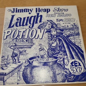 May include: Vintage vinyl record album cover for "The Jimmy Heap Show" featuring Ken Idaho. The cover art is a blue and white illustration of a witch stirring a cauldron labeled "Laugh Potion". A cat and owl are also featured.