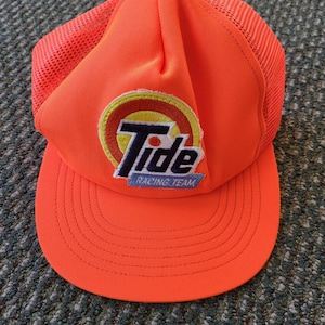Könnte beinhalten: Leuchtend orangefarbene Trucker-Cap mit Mesh-Rückseite und gebogenem Schirm. Auf der Vorderseite befindet sich das Tide-Logo und der Text "Racing Team". Ein auffälliges Accessoire.