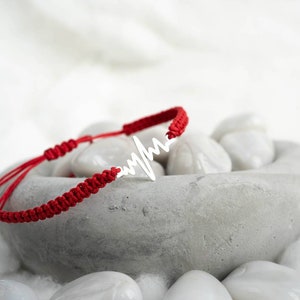 May include: Red string bracelet with a silver heart beat charm. The bracelet is lying on a bed of white stones.