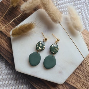 May include: A pair of green and gold dangle earrings with a small round green stone and a larger green stone with gold flecks. The earrings are on a white surface.