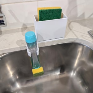 May include: A white rectangular sponge holder containing a yellow and green sponge, positioned next to a dish wand with a light blue cap and a yellow and green sponge head, which is attached to the side of a stainless steel kitchen sink.