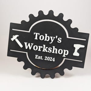 May include: Black wooden sign with a gear shape and the text "Toby's Workshop Est. 2024" in white lettering. A hammer and drill icon are on either side of the text.