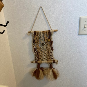 May include: A macrame wall hanging with brown, beige, and white yarn. The hanging has wooden dowels at the top and bottom and features brown and beige tassels.