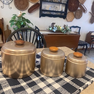 May include: Set of three vintage copper-toned canisters with wooden lids. The canisters are cylindrical and vary in size, with the largest one on the left. They sit on a blue and white checkered tablecloth, suggesting kitchen storage.