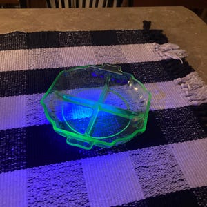 May include: A translucent, light green, four-sectioned glass serving dish with a scalloped edge and two small handles. The glass glows with a bright neon green hue under ultraviolet light, highlighting its segmented design and smooth surface.