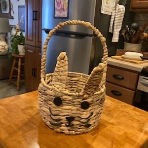 May include: A woven basket in the shape of a bunny with a handle. The basket is made of natural-colored woven material with black accents for the eyes and nose. The basket has two upright ear shapes.
