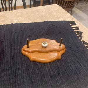 May include: A wooden decorative plate holder with a light brown finish. It features a white and gold knob in the center and two brass posts. The holder is placed on a dark blue textured mat.