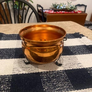 May include: A round, metallic copper-colored bowl with a smooth, reflective finish. The bowl features a wide, open top and is supported by three small, curved black metal legs attached to the base, giving it a distinct, elevated, cauldron-like appearance.