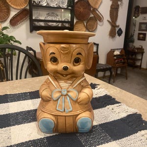 May include: A vintage ceramic cookie jar shaped like a cartoon dog wearing a sailor hat and uniform. The jar is primarily tan with blue accents on the bow tie and feet. The lid sits on top of the dog's head. The jar is sitting on a blue and white checkered table runner.