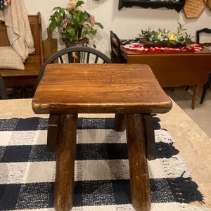 May include: A small, square wooden stool with a dark brown, weathered finish. The stool features a flat, rectangular top surface with visible wood grain and natural imperfections, supported by four thick, sturdy, cylindrical wooden legs.