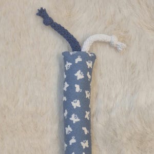 Könnte beinhalten: Ein zylindrisches Katzenspielzeug aus blauem Stoff mit weißem Schmetterlingsmuster. Es hat zwei seilartige Aufsätze, einen blauen und einen weißen, die an den Enden verknotet sind. Für Katzen zum Spielen.