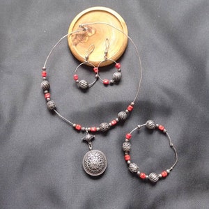 Puede incluir: Un collar de plata con un colgante, pendientes y una pulsera. El collar y la pulsera tienen cuentas rojas y detalles plateados. El colgante es de diseño redondo y adornado.