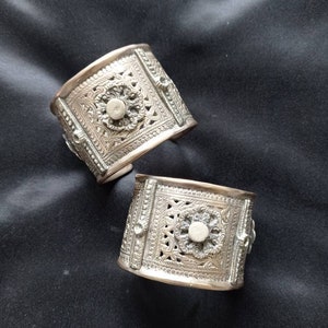 May include: Two silver bracelets with intricate floral designs and a circular centre piece. The bracelets are on a black fabric background.