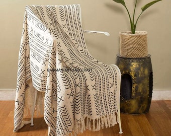 May include: A white throw blanket with black geometric patterns and fringe. The blanket is draped over a white chair and a small plant is in a woven basket on a gold metal stool.