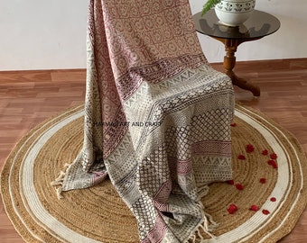 Puede incluir: Una alfombra redonda de yute con ribete blanco y una manta estampada drapeada sobre una silla. La alfombra tiene un tono natural y terroso y la manta presenta un diseño geométrico en tonos rosa, gris y negro.
