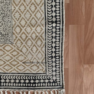 Puede incluir: Una alfombra beige y negra con estampado y borde de flecos. La alfombra tiene un dise&ntilde;o geom&eacute;trico con detalles en blanco y negro. El texto "HARMAN ART AND CRAFT" es visible en la alfombra.