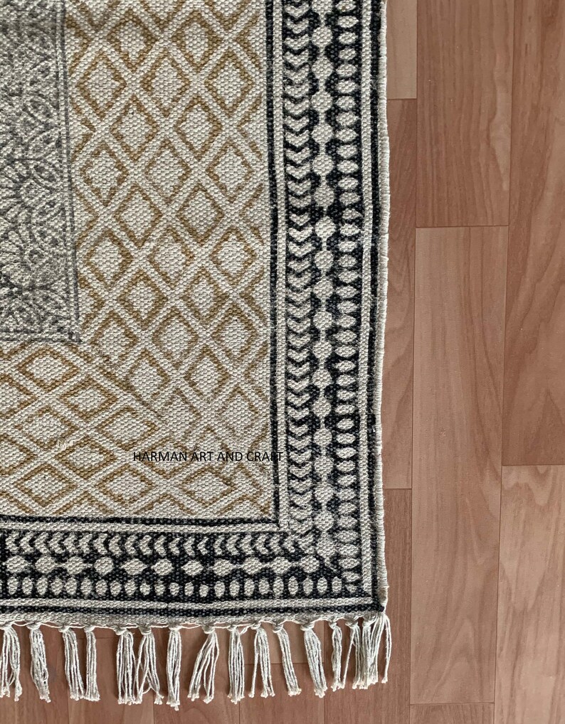 Puede incluir: Una alfombra beige y negra con estampado y borde de flecos. La alfombra tiene un dise&ntilde;o geom&eacute;trico con detalles en blanco y negro. El texto "HARMAN ART AND CRAFT" es visible en la alfombra.