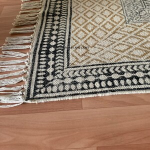 Puede incluir: Una alfombra blanca y negra con un borde de flecos. La alfombra tiene un dise&ntilde;o geom&eacute;trico con detalles en blanco y negro.