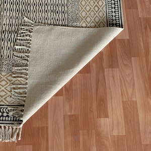 Puede incluir: Una alfombra blanca y negra con estampado y borde de flecos. La alfombra est&aacute; doblada por la mitad y se ve la parte inferior. La alfombra est&aacute; sobre un suelo de madera marr&oacute;n claro.