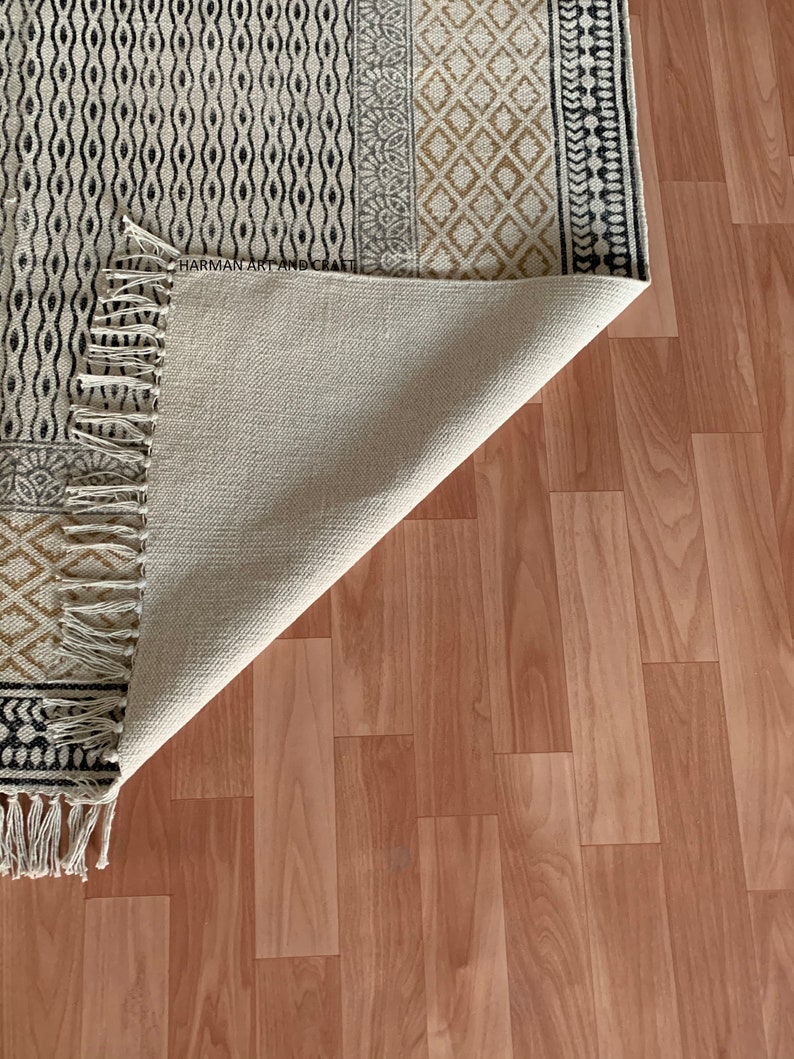 Puede incluir: Una alfombra blanca y negra con estampado y borde de flecos. La alfombra est&aacute; doblada por la mitad y se ve la parte inferior. La alfombra est&aacute; sobre un suelo de madera marr&oacute;n claro.