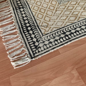Puede incluir: Una alfombra blanca y negra con un borde de flecos. La alfombra tiene un dise&ntilde;o geom&eacute;trico con diamantes blancos y negros y un borde marr&oacute;n y blanco.