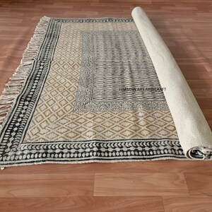 Puede incluir: Una alfombra de algod&oacute;n tejida a mano con un patr&oacute;n geom&eacute;trico en blanco y negro y un borde beige. La alfombra tiene flecos en todos los lados y est&aacute; enrollada.