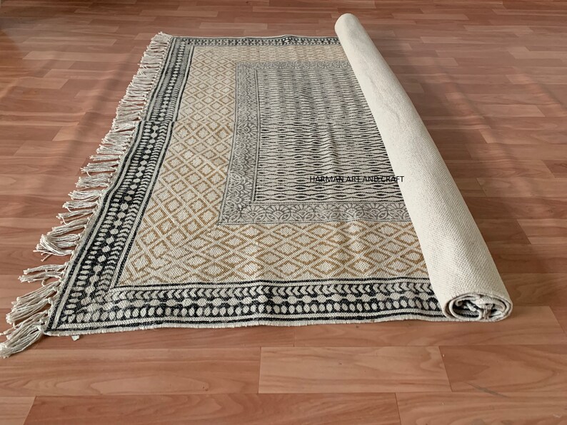 Puede incluir: Una alfombra de algod&oacute;n tejida a mano con un patr&oacute;n geom&eacute;trico en blanco y negro y un borde beige. La alfombra tiene flecos en todos los lados y est&aacute; enrollada.