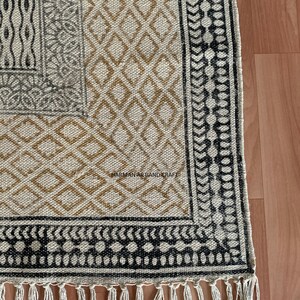 Puede incluir: Una alfombra beige y marr&oacute;n con estampado y ribete negro y blanco. La alfombra tiene un borde de flecos y el texto "HARMAN ART AND CRAFT" es visible en la esquina inferior derecha.