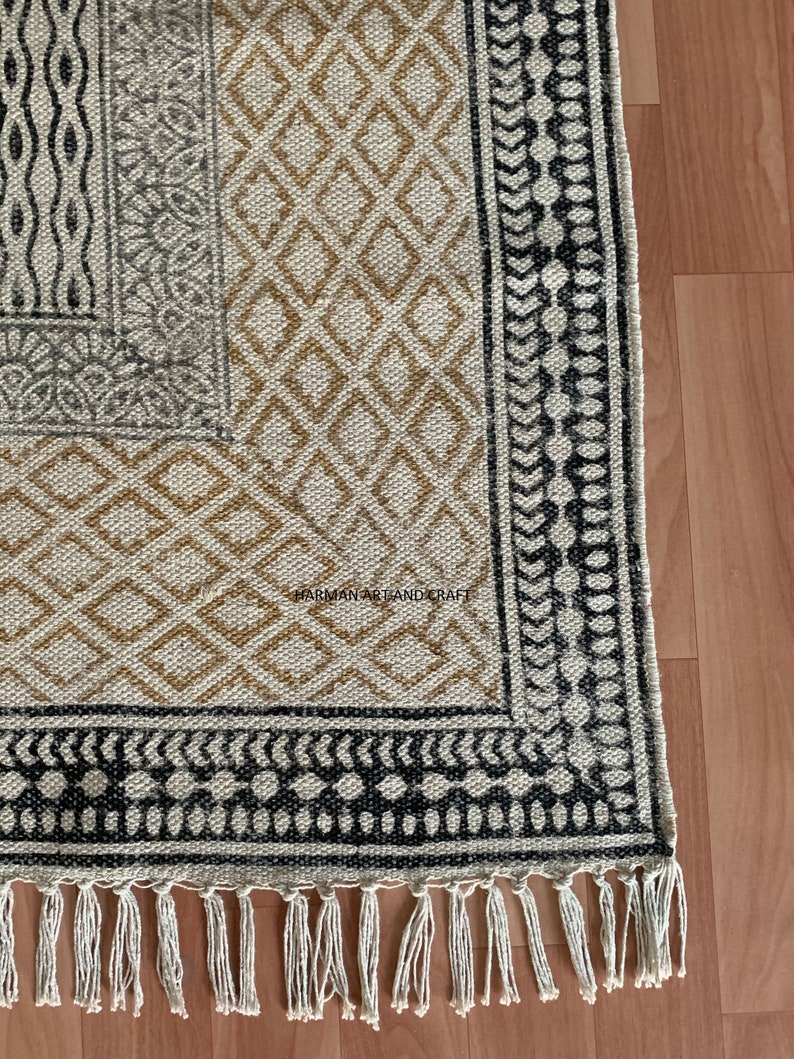 Puede incluir: Una alfombra beige y marr&oacute;n con estampado y ribete negro y blanco. La alfombra tiene un borde de flecos y el texto "HARMAN ART AND CRAFT" es visible en la esquina inferior derecha.